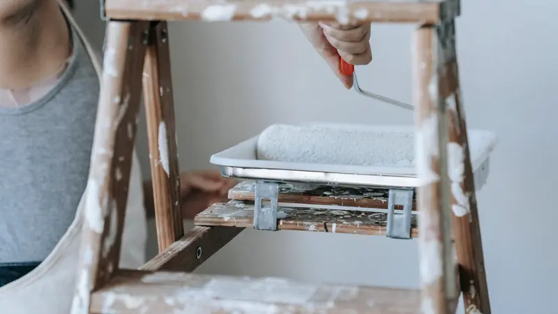 Personne au pied d'un escabeau taché de peinture, rouleau en main au-dessus d'un bac de peinture blanche