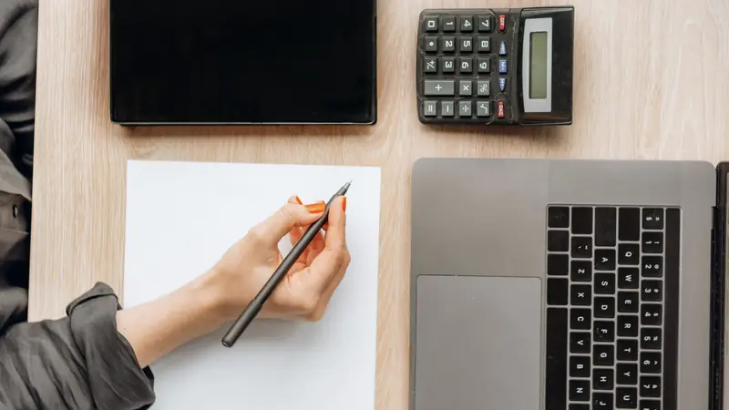 Main prenant des notes à côté d'une calculatrice et d'un ordinateur portable