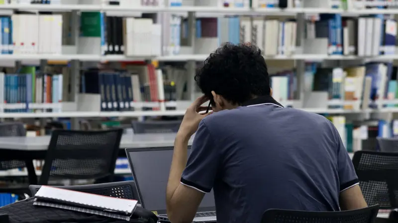 Étudiant vu de dos, travaillant sur un ordinateur portable dans une bibliothèque, un cahier posé à côté