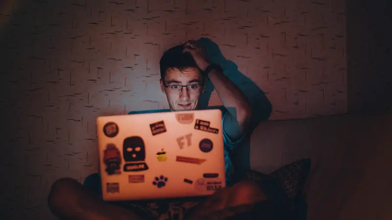 Jeune homme à lunettes, la main dans les cheveux, devant un ordinateur portable couvert d'autocollants dans une pièce sombre