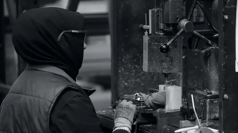 Ouvrière en lunettes de protection et gants perçant une pièce métallique sur une perceuse à colonne, photo en noir et blanc
