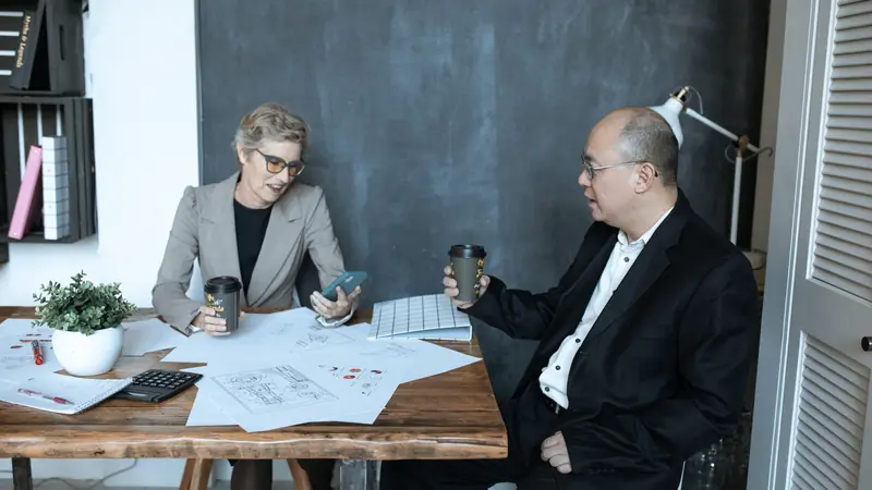 Deux professionnels en veste discutant autour d'une table couverte de plans, d'une calculatrice et de gobelets de café