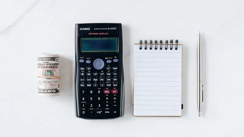 Calculatrice, rouleau de billets, carnet et stylo alignés sur une table blanche