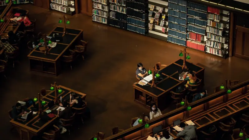 Salle de lecture d'une bibliothèque vue d'en haut, étudiants travaillant sur des ordinateurs portables sous des lampes vertes
