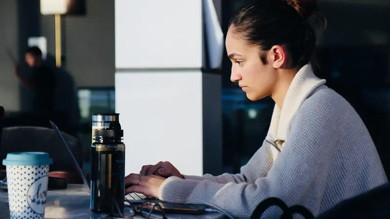 Jeune femme de profil tapant sur un ordinateur portable, une gourde et un gobelet posés sur la table
