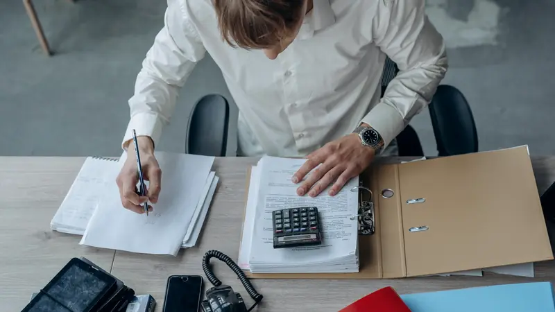 Homme en chemise blanche annotant des feuilles à son bureau, une calculatrice posée sur une pile de documents dans un classeur ouvert