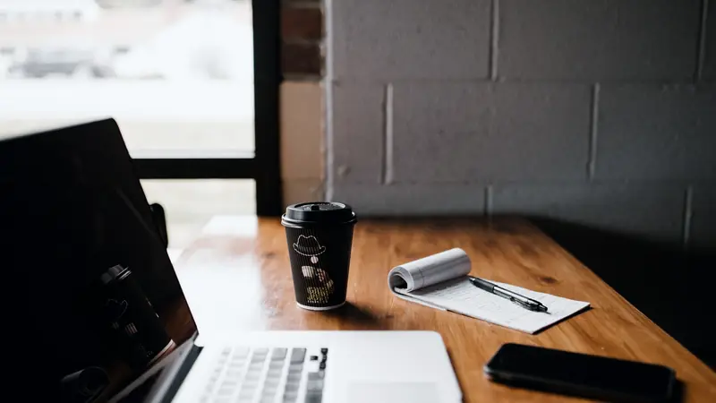 Ordinateur portable, café à emporter, bloc-notes et téléphone sur un bureau en bois près d'une fenêtre