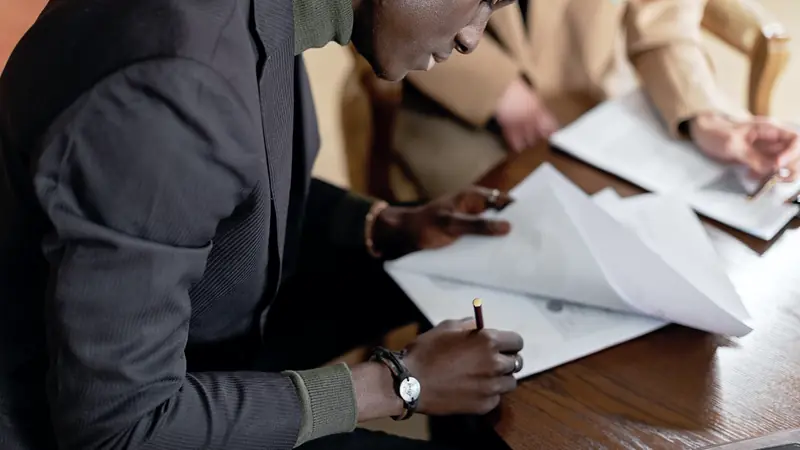 Homme signant des documents posés sur une table en bois, une autre personne relisant un dossier en face