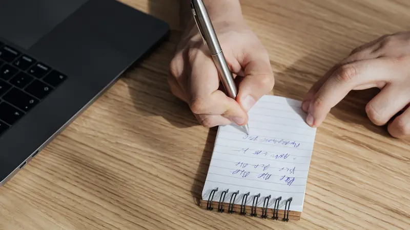 Main écrivant au stylo sur un petit carnet à spirale, à côté d'un ordinateur portable