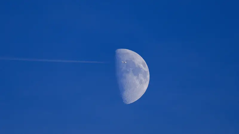 Avion passant devant une demi-lune dans un ciel bleu, traînée blanche derrière lui