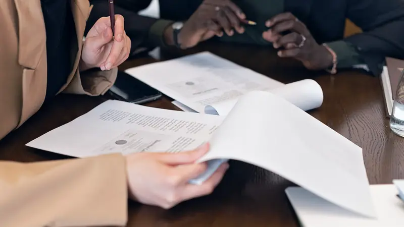 Mains de deux personnes relisant des documents imprimés sur une table en bois, stylo en main