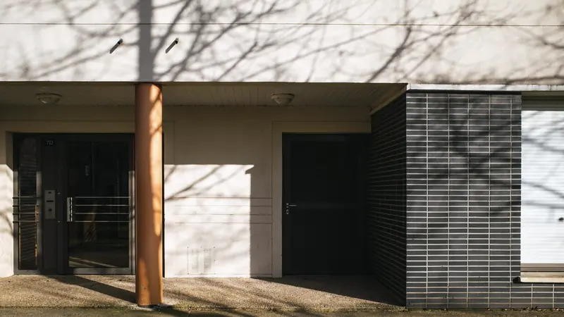 Entrée vitrée d'un immeuble résidentiel moderne, pilier orange et ombres d'arbres sur la façade