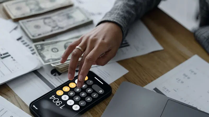 Main qui tape un calcul sur la calculatrice d'un smartphone, entre des liasses de billets et des documents chiffrés