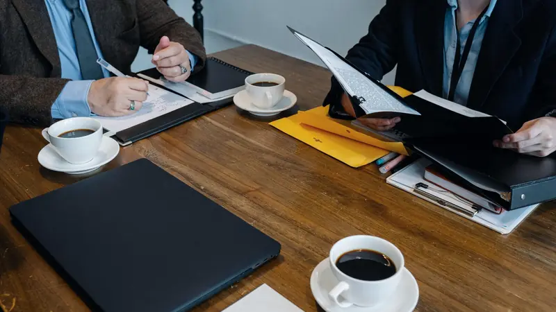 Deux personnes en tenue de bureau autour d'une table en bois, avec des dossiers, un carnet de notes et des tasses de café
