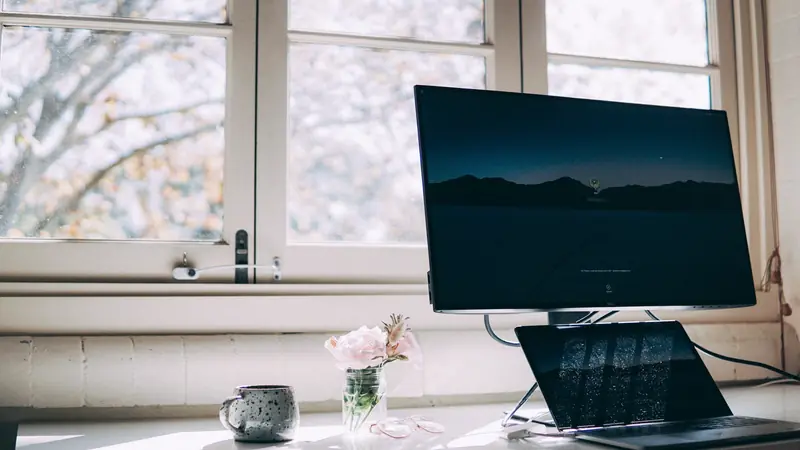 Bureau installé devant une fenêtre avec un écran, un ordinateur portable et un petit bouquet