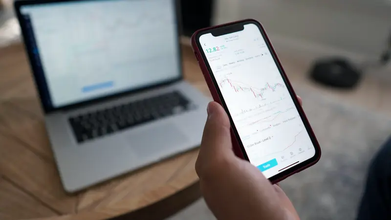 Main tenant un smartphone affichant une application de suivi financier devant un ordinateur portable