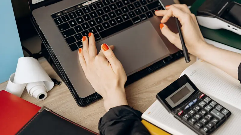 Mains aux ongles orange sur le pavé d'un ordinateur portable ouvert sur un tableur, à côté d'une calculatrice, d'un carnet quadrillé et d'un rouleau de papier de caisse