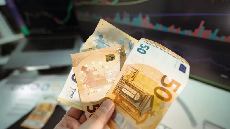 Main tenant des billets de 50 et 20 euros devant un écran affichant des graphiques financiers