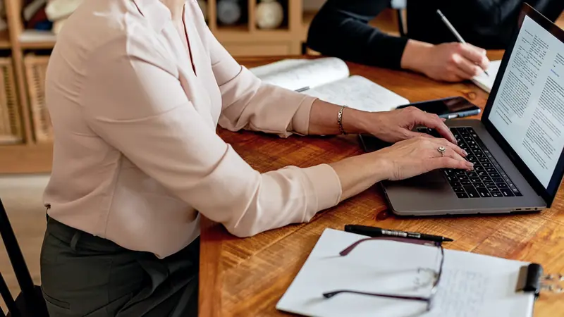 Femme tapant sur un ordinateur portable à une table en bois, un carnet de notes et des lunettes posés à côté
