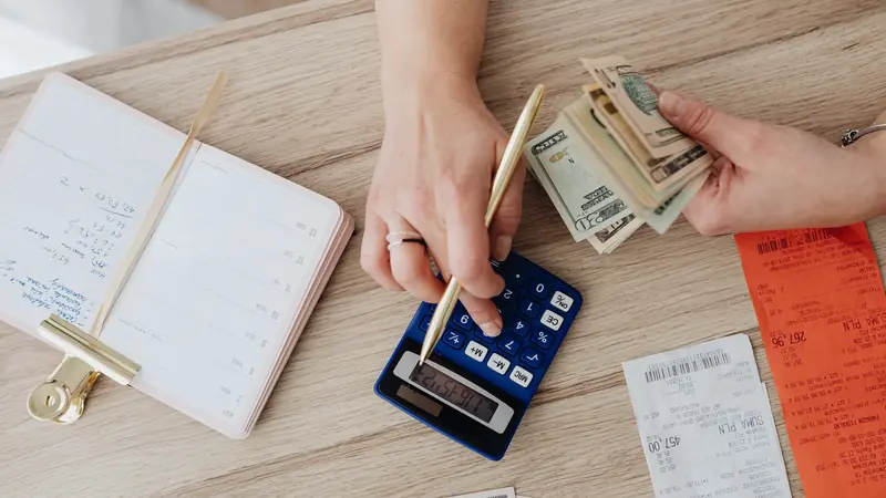 Mains tapant sur une calculatrice bleue avec un stylo doré, à côté de tickets de caisse, de billets et d'un agenda ouvert