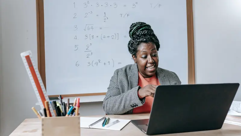 Enseignante assise à son bureau devant un ordinateur portable, un tableau blanc couvert d'exercices de calcul derrière elle