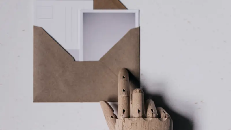 Main en bois articulée tenant une enveloppe kraft ouverte qui contient deux cartes blanches