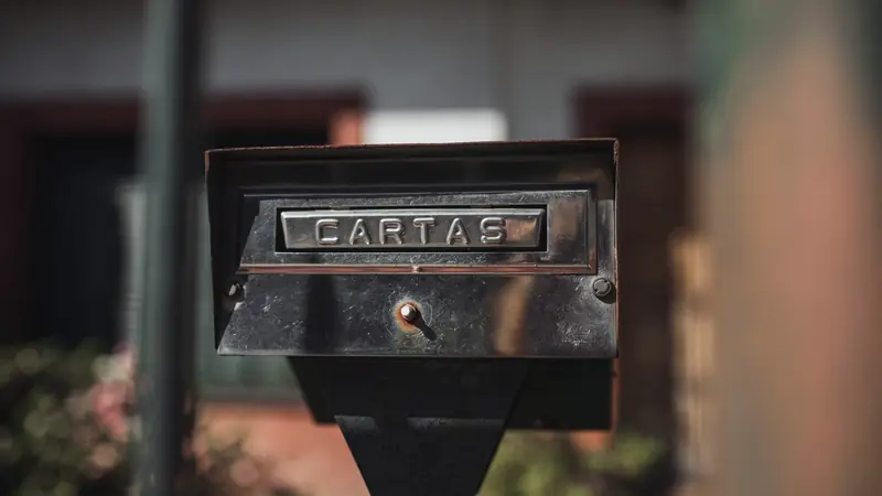 Boîte aux lettres métallique sur pied, avec une fente gravée « Cartas », devant une maison floue
